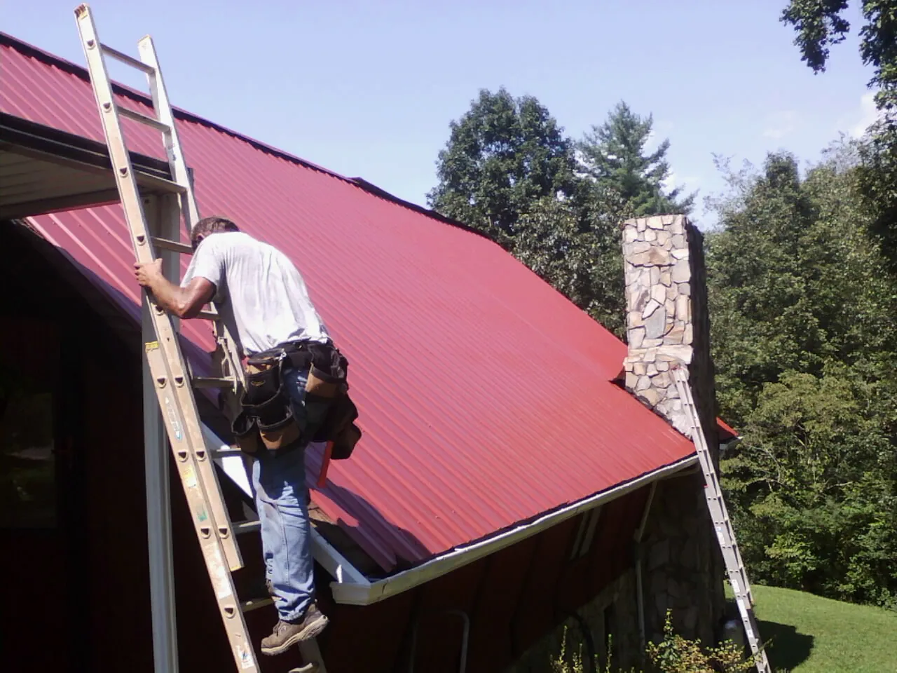 Roofer on a ladder beside a red metal roof and stone chimney — Soffit & Fascia Repair by Roofing Brothers in North Easton
